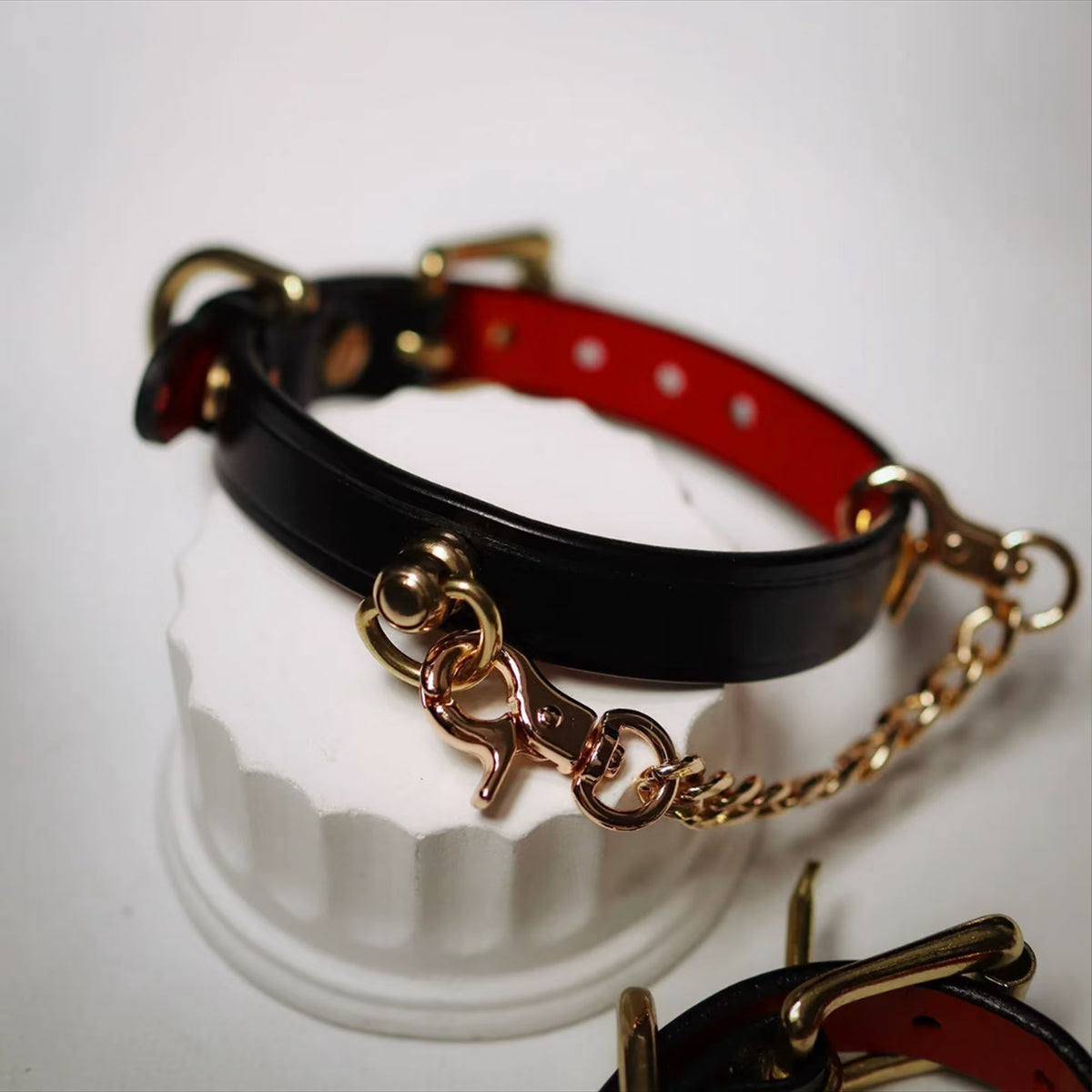Black leather BDSM collar on white display stand – red lining gold hardware chain leash D-ring buckle detail
