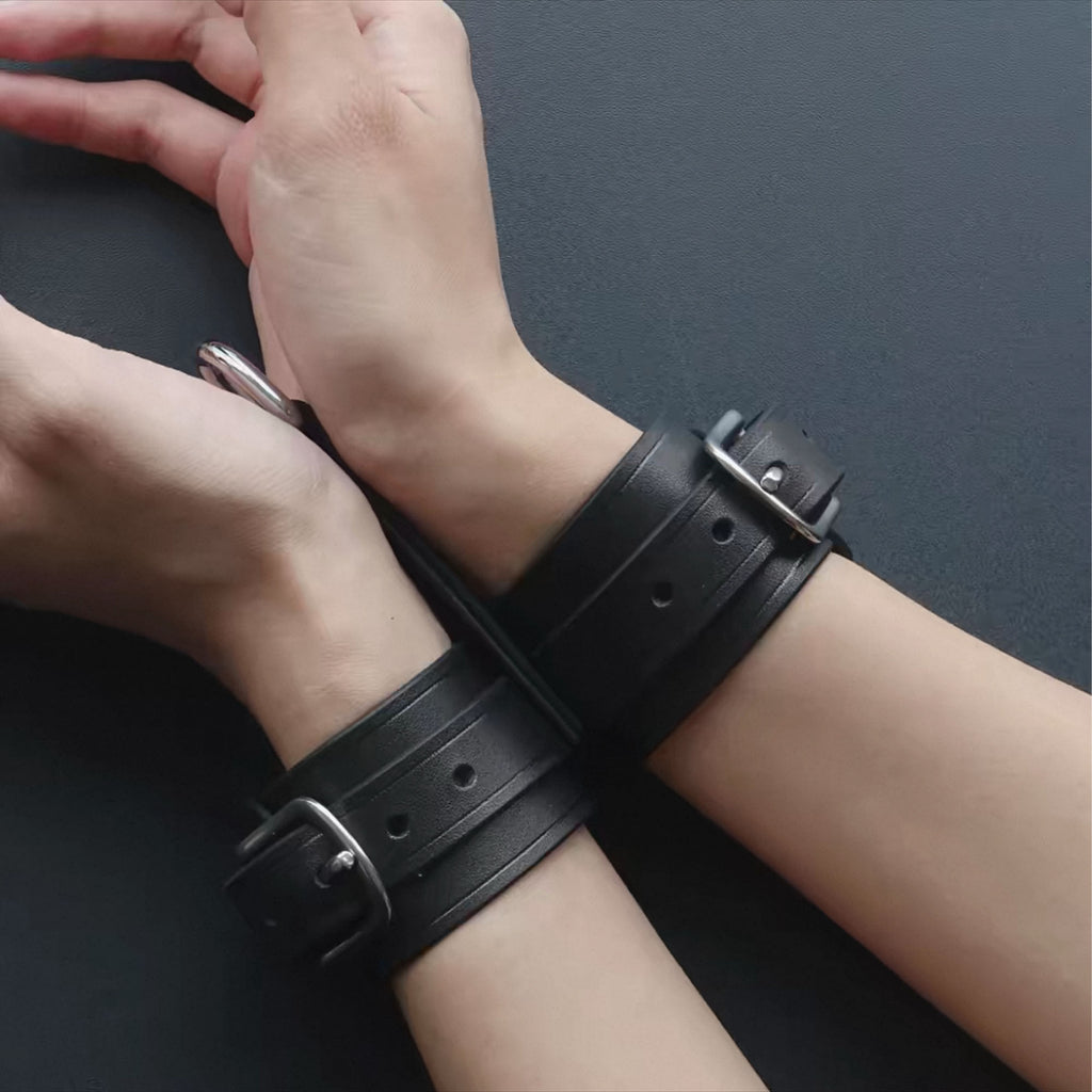 Black leather wrist cuffs worn on model crossed wrists overhead – silver buckle D-ring BDSM restraint close-up
