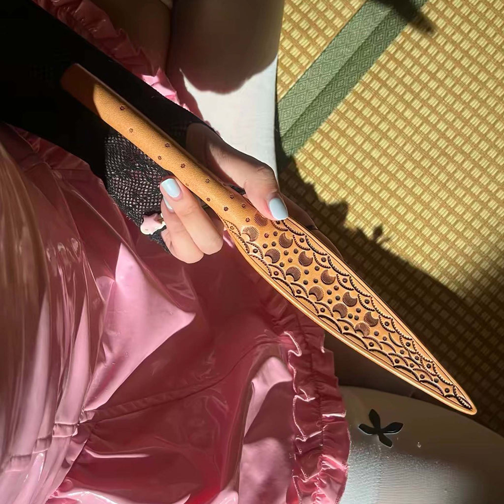Natural tooled leather paddle gripped in hand – carved floral blade pink satin background BDSM lifestyle editorial
