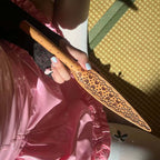 Natural tooled leather paddle gripped in hand – carved floral blade pink satin background BDSM lifestyle editorial
