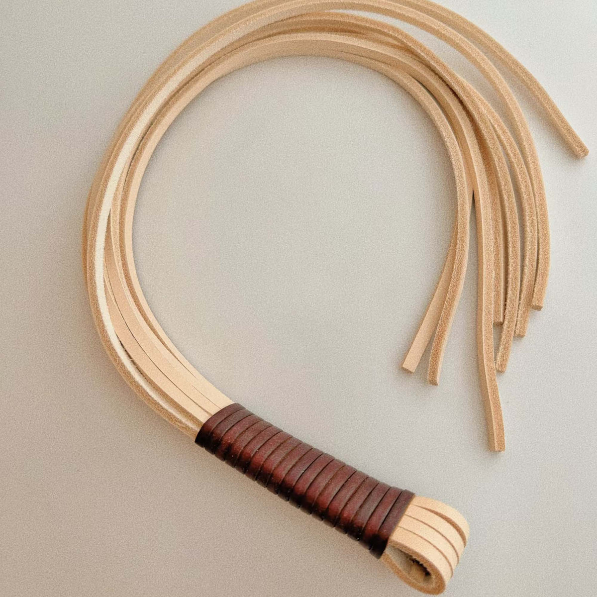 Natural vegetable tanned leather flogger coiled on light gray surface – cream tails looped dark brown wrapped handle full view BDSM
