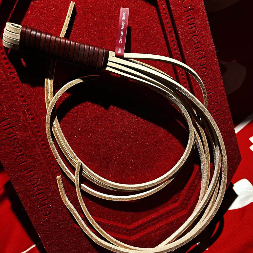Natural leather flogger coiled on red velvet surface with brand label – cream tails dark brown handle Chuncui Design Studio BDSM handmade
