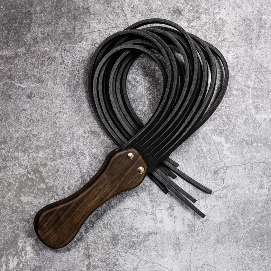Black leather flogger coiled on stone surface – walnut wood handle gold rivet tails looped BDSM product detail
