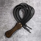 Black leather flogger coiled on stone surface – walnut wood handle gold rivet tails looped BDSM product detail
