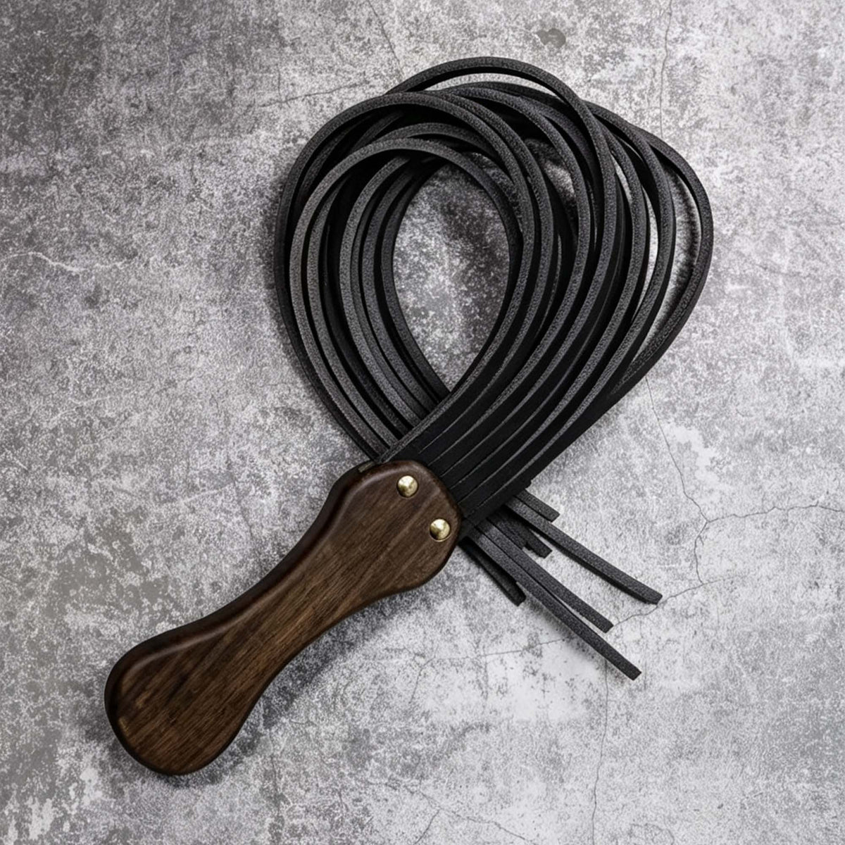Black leather flogger coiled on stone surface – walnut wood handle gold rivet tails looped BDSM product detail
