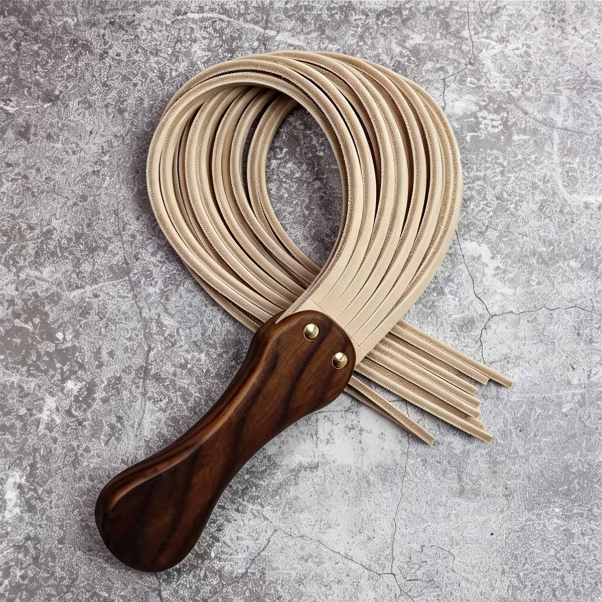 Natural tan leather flogger coiled on stone surface – walnut wood handle gold rivet tails looped BDSM product detail
