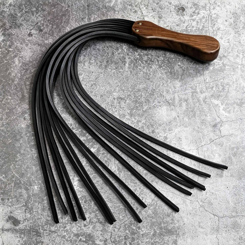 Black leather flogger flat lay product display – walnut wood handle gold rivet vegetable tanned multiple tails BDSM handmade ShadowedBodies
