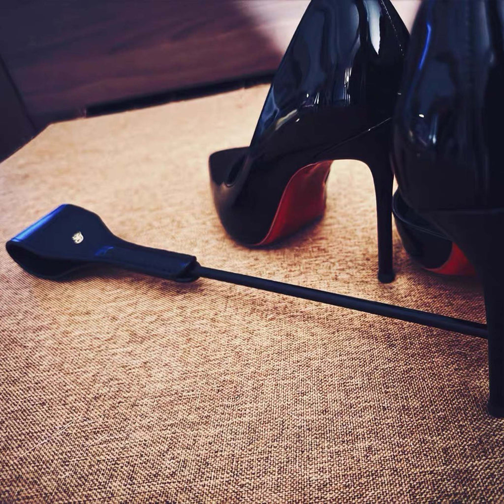 Black leather riding crop on floor in front of black high heels – lifestyle editorial BDSM dominance scene
