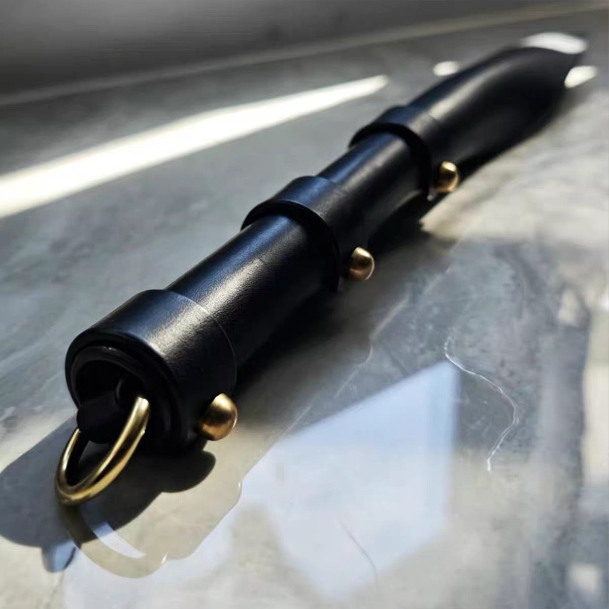 Black leather soft paddle handle close-up on marble surface – segmented wrap gold rivet D-ring detail cowhide BDSM
