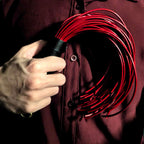 Red leather whip gripped in hand against dark shirt – black handle red lash coil BDSM impact play editorial
