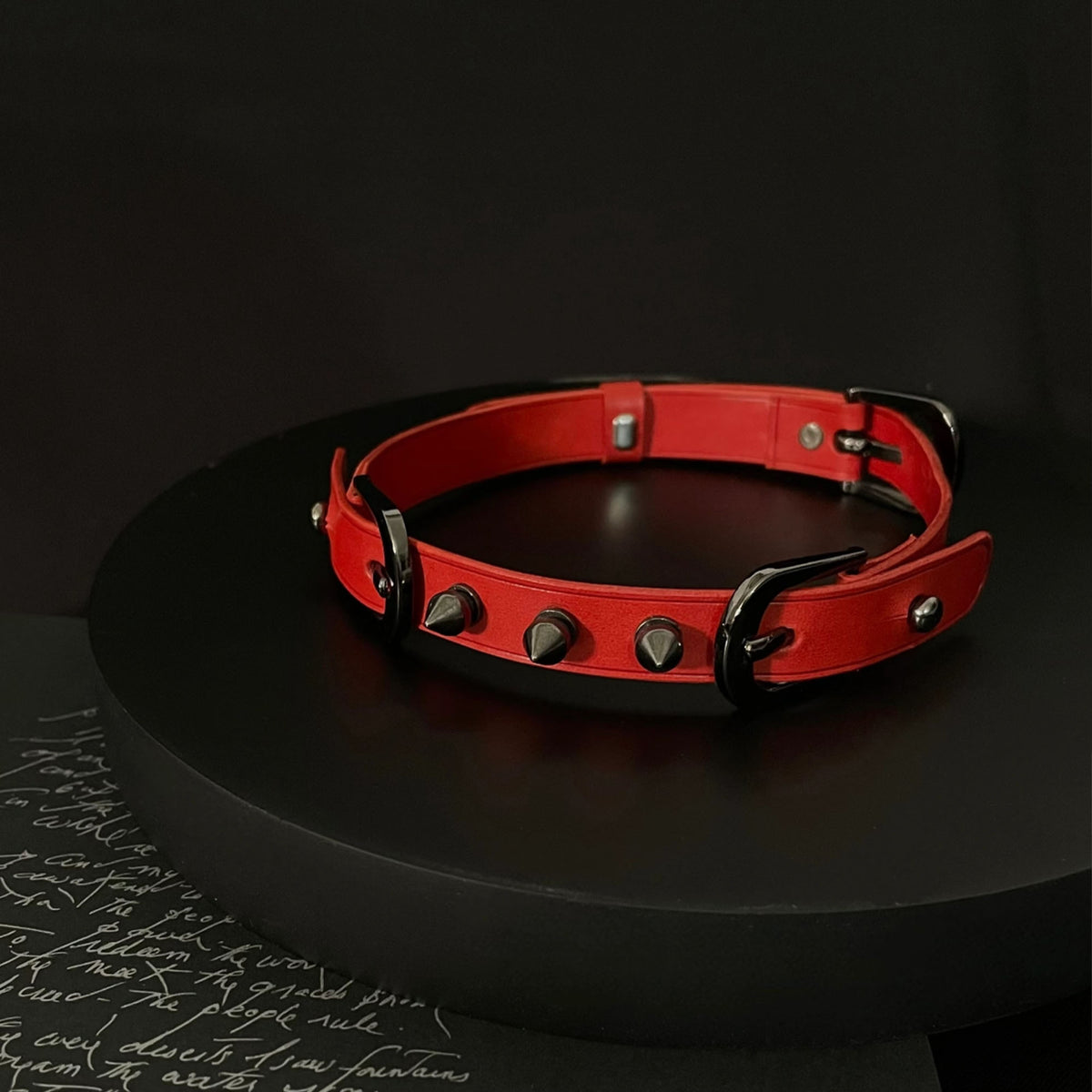 Matte red leather BDSM choker on black display stand – black cone spike rivets double buckle dark editorial
