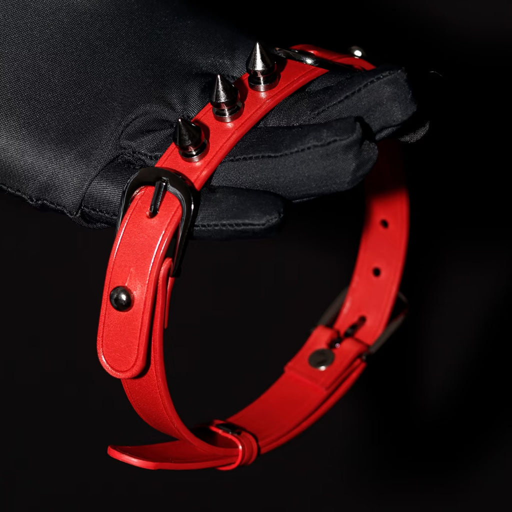 Matte red leather spiked choker and wrist cuff held in black gloved hands – silver cone spikes black buckle detail
