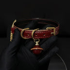 Burgundy leather BDSM collar held in black gloved hands – gold square buckle D-ring red bell pendant chain detail
