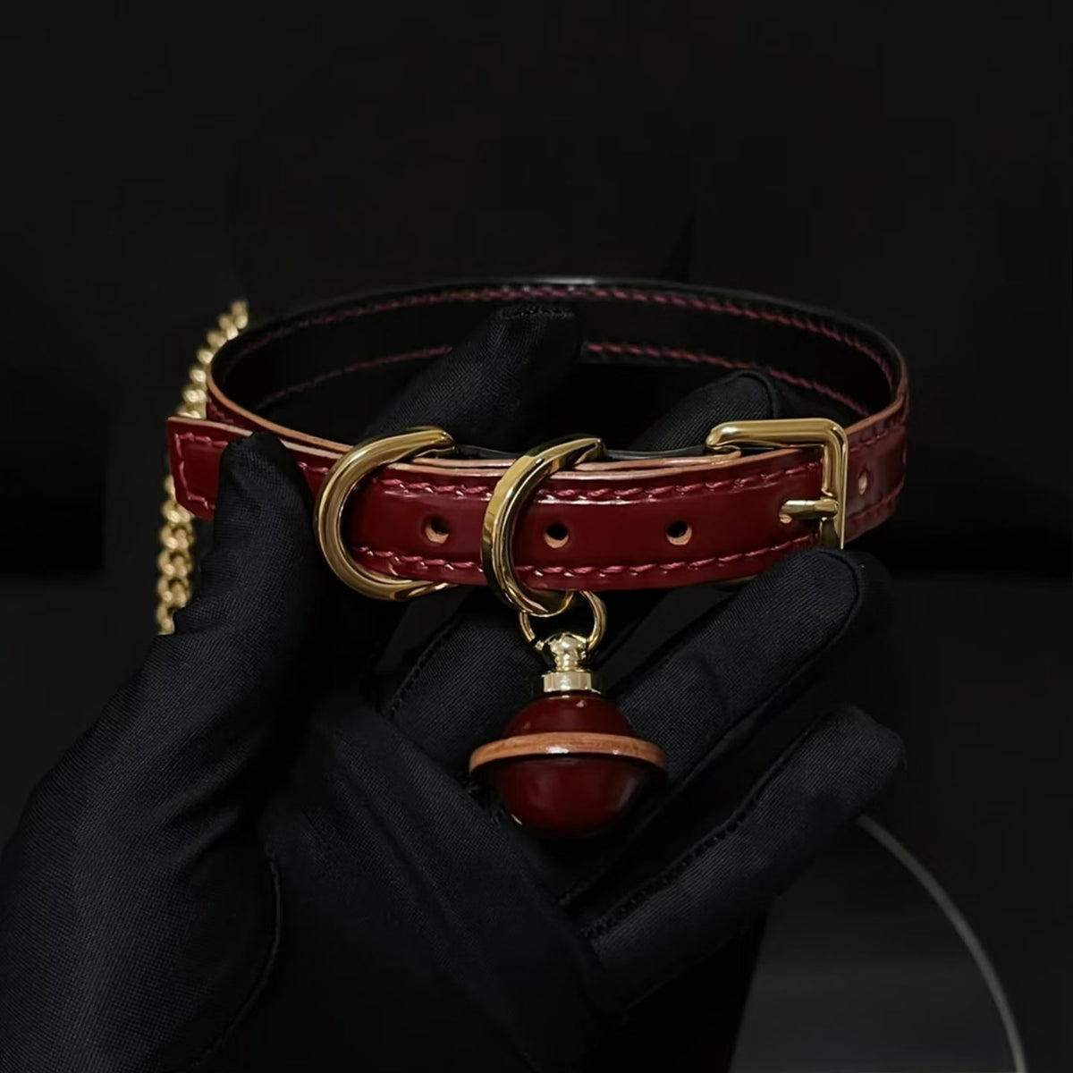 Burgundy leather BDSM collar held in black gloved hands – gold square buckle D-ring red bell pendant chain detail
