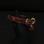 Burgundy leather BDSM collar angled view – gold buckle D-ring red bell pendant saddle stitching detail
