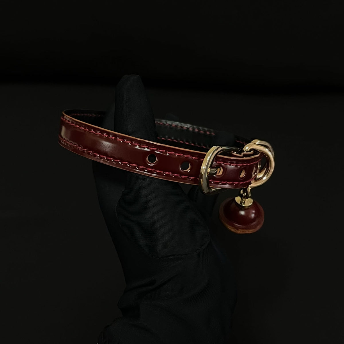 Burgundy leather BDSM collar angled view – gold buckle D-ring red bell pendant saddle stitching detail
