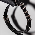Matte black leather spiked BDSM choker held in black gloved hands – silver cone spikes double buckle D-ring close-up
