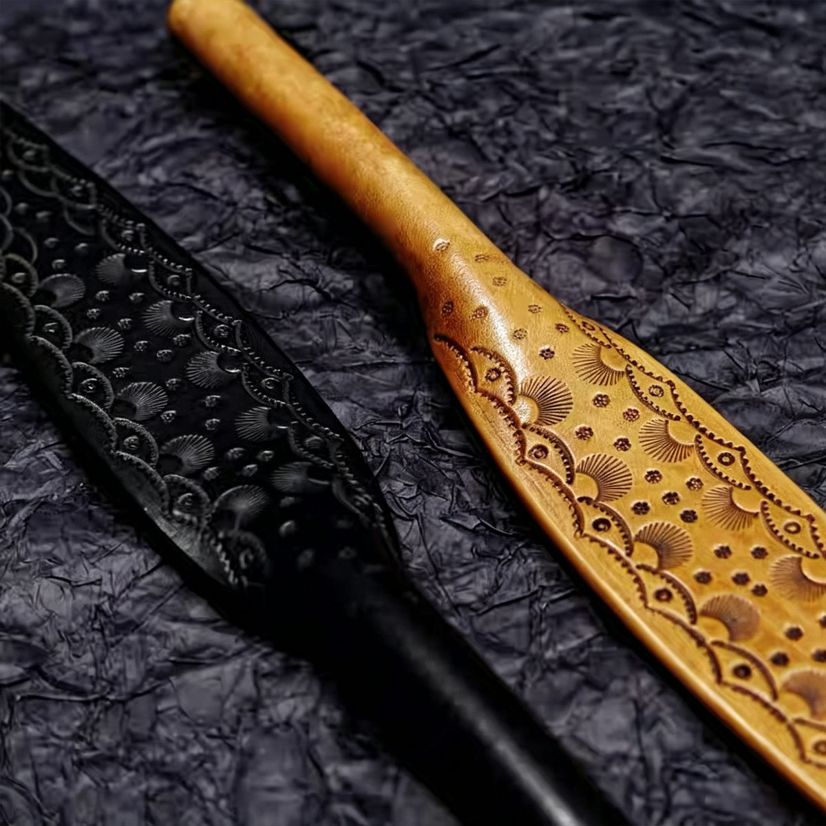Black and natural tooled leather paddles side by side on dark textured surface – carved pattern contrast close-up BDSM handmade
