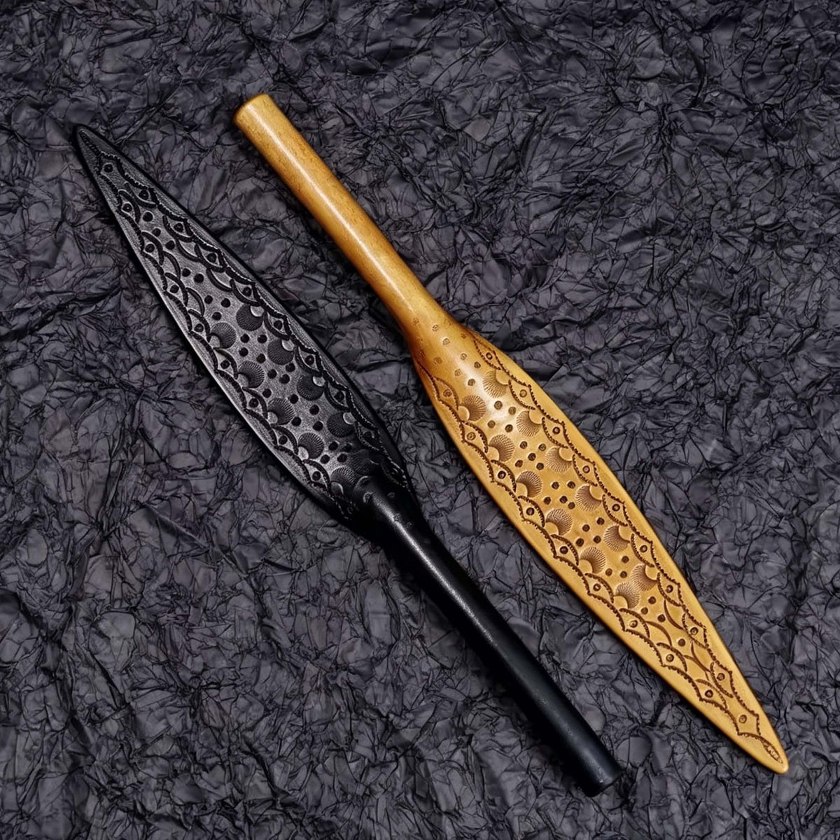 Black plain and natural tooled leather paddles flat lay on dark textured surface – both styles full view leaf blade BDSM comparison
